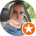 A person is seated in a car, looking at the camera with a neutral facial expression. Each reflects outside the car window. A white star on an orange circle is superimposed on the lower right of the image, reminiscent of a modern web design footer icon.
