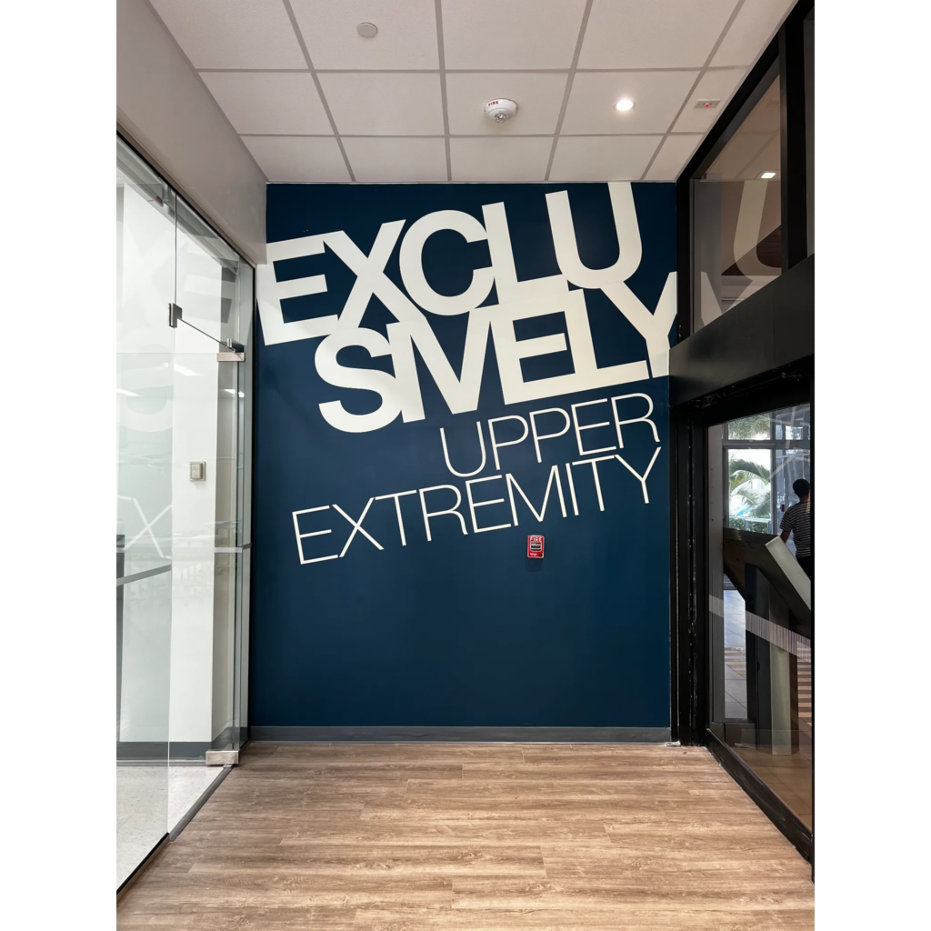 A hallway with a blue wall featuring large, stylized white text reading "EXCLUSIVELY UPPER EXTREMITY." Adjacent to a glass door on the left and black-framed windows on the right.