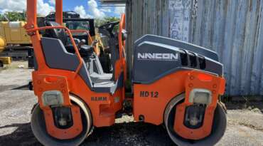 An orange and gray HAMM HD12 road roller with "NANCON ENGINEERING" branding and custom fleet wraps is parked on gravel beside a weathered shipping container.