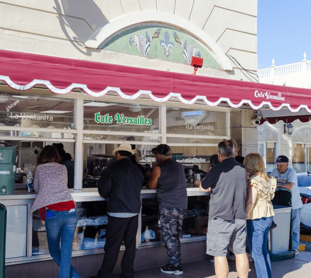 9c19ba81ec9c37e54eeb738a3a2672db356a507bc300204f4f2e94e88e087767 People stand in line at an outdoor window counter of Café Versailles in Miami, beneath a red awning and a mosaic sign, waiting for authentic Cuban coffee.